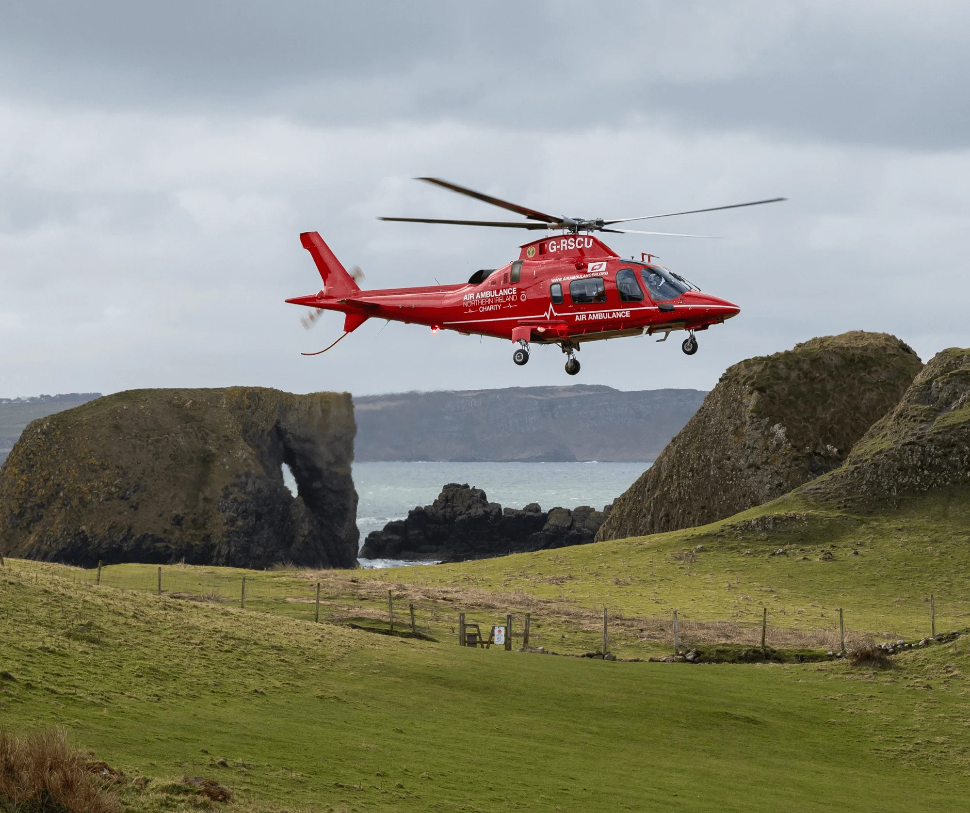 An Air Ambulance NI helicopter hovers above grassy cliffs by the sea on a cloudy day.