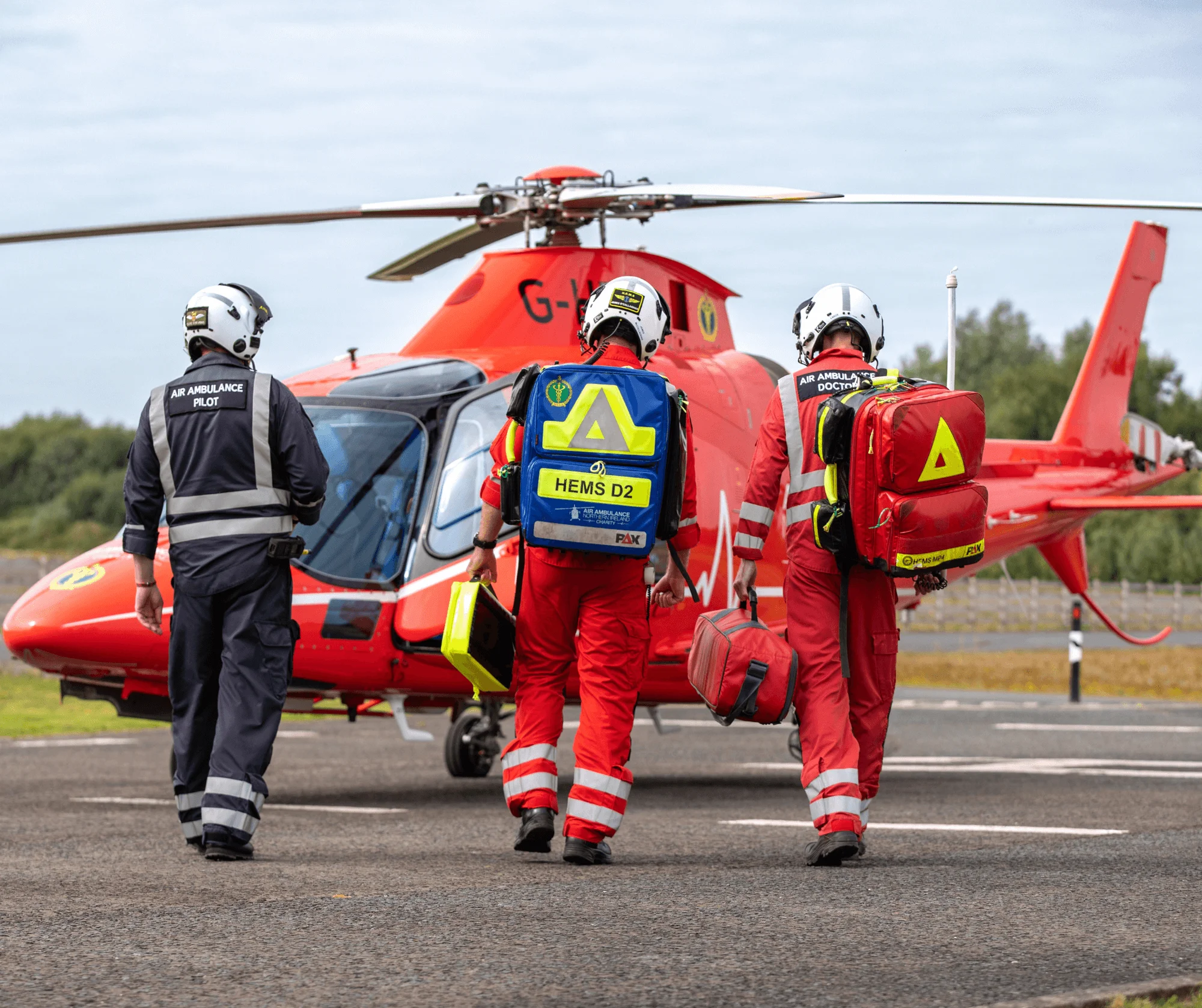 Air Ambulance NI crew in red uniforms and helmets walk toward a red helicopter on a helipad.